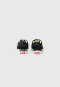 Schwarze Wildleder Slipper mit einer weißen Gummisohle und rotem Vans-Logo. Kontrastnähte und elastische seitliche Akzente für ein bequemes Anziehen.