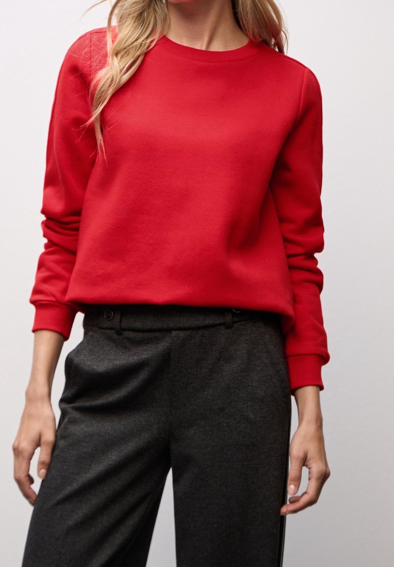 Personne portant un sweat-shirt rouge vif à manches longues et un pantalon droit gris foncé sur un fond uni.