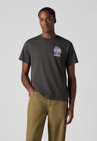 Gray cotton t-shirt featuring a colorful graphic print on the left chest, paired with light brown pants; short sleeves and relaxed fit.