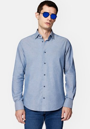 Jasnoniebieska koszula z długim rękawem, z niebieskimi guzikami i miękką fakturą, w połączeniu z okrągłymi niebieskimi okularami przeciwsłonecznymi.