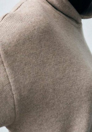 Béžový ribbed rolák s jemnou pletenou texturou a přiléhavým střihem, zobrazený od ramene dolů, s vysokým límcem.