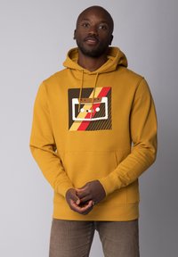 Sweat à capuche jaune avec poche frontale et cordon de serrage. Présente un design graphique avec une cassette, des rayures rouges, marron et jaunes.