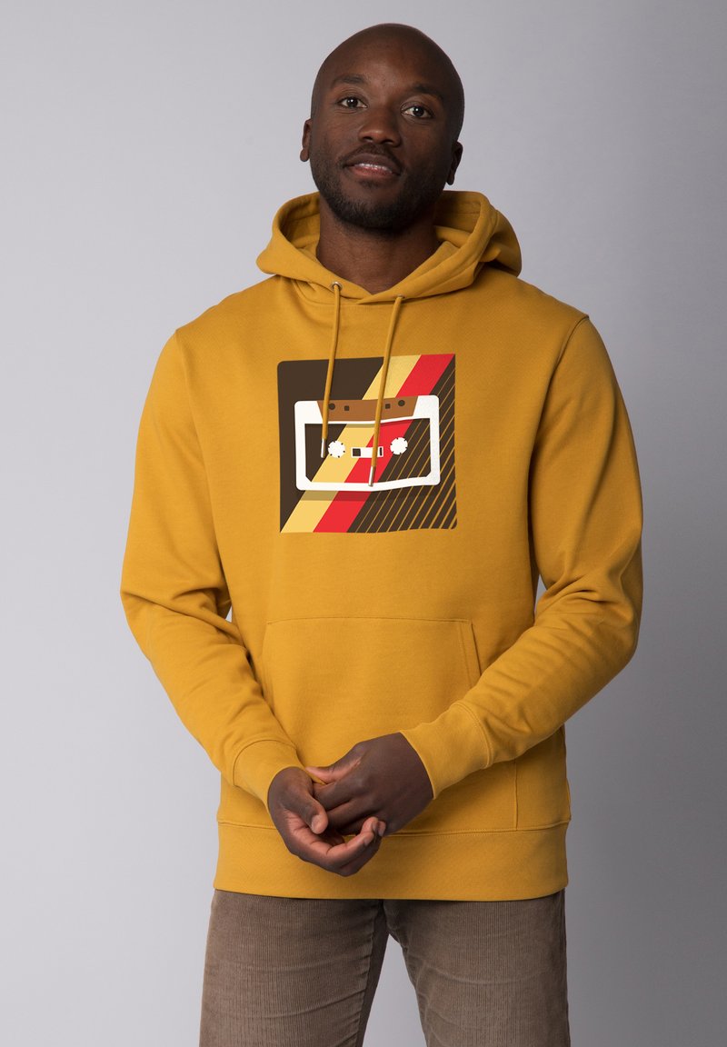 Sweat à capuche jaune avec poche frontale et cordon de serrage. Présente un design graphique avec une cassette, des rayures rouges, marron et jaunes.