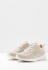Un paio di sneaker lavorate a maglia beige con suole e lacci bianchi, esposte angolate a destra su una superficie bianca contro uno sfondo semplice.