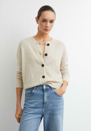 Frau mit braunen, nach hinten gebundenen Haaren, die eine cremefarbene geknöpfte Strickjacke und hellblaue High-Waist-Jeans trägt, steht vor einem weißen Hintergrund.