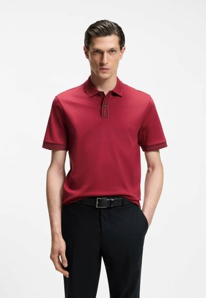 Man with slicked-back hair wearing a red short-sleeve polo shirt and black pants, standing with one hand in pocket against white background.