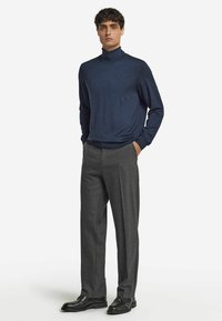 En man står med händerna i fickorna, klädd i en marinblå polotröja, grå kostymbyxor och svarta läderskor.