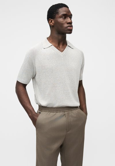 Ελαφρώς γκρι κοντομάνικη πλεκτή πόλο με V-λαιμόκοψη και επίπεδη γιακά, συνδυασμένη με καφέ στενά παντελόνια με ελαστική μέση.