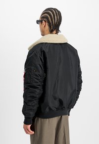 Cazadora bomber negra con un cuello de piel de oveja en color crema, puños acanalados y detalles fruncidos en las mangas. Presenta un bolsillo lateral con detalle de herrajes.