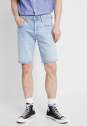 Man wearing light blue denim shorts, black high-top sneakers with white laces, white socks, and a blue shirt with one hand in pocket.