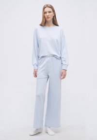 Γυναίκα που στέκεται φορώντας ανοιχτό μπλε μπλούζα με μακριά μανίκια και φαρδιά παντελόνια με ζώνη μέσης "POLO RALPH LAUREN" σε απλό λευκό φόντο.