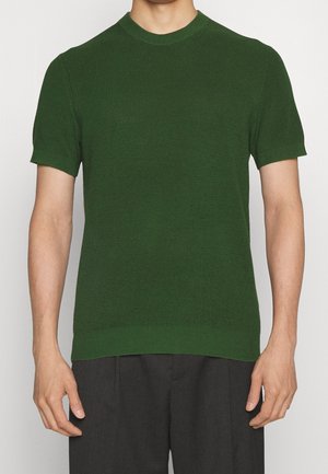 Mann trägt einen olivgrünen, kurzärmeligen Pullover mit strukturierter Strickoptik und Rundhalsausschnitt sowie dunkelgraue Hosen.