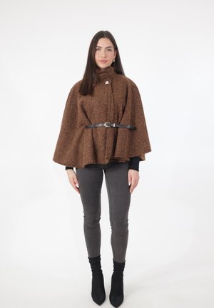 Femme portant une cape marron en laine avec une ceinture noire, un haut à manches longues noir, un jean skinny gris et des bottines noires sur fond blanc.