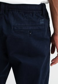 Marineblauwe broek met een elastische tailleband, een achterzak met knoopsluiting en een klein merkembleem boven de zak.