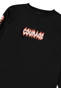 Fekete hosszúujjú póló, amelyen a "COURAGE" felirat látható lángmintában, narancssárga és piros árnyalatban, átkelve a mellkason és végig a ujjak mentén.