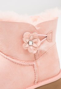 Botte en daim rose doux avec doublure moelleuse, ornée d'une décoration en cuir en forme de fleur avec une gemme transparente centrale sur le côté.