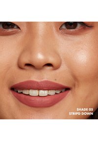 Primo piano di labbra sorridenti con rossetto opaco di colore rosa-marrone chiaro. Carnagione naturale e guance in evidenza. Il testo in basso indica la tonalità.