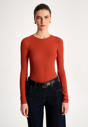 Kobieta z zaczesanymi do tyłu włosami nosi dopasowaną bluzkę w kolorze rdzawego pomarańczu z długim rękawem, ciemne jeansy z wysokim stanem i czarny pasek z klamrą.