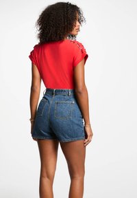 Rød t-skjorte med korte ermer og metallhulldetaljer, kombinert med høyt midje blå denimshorts med to baklommer og rå kant.