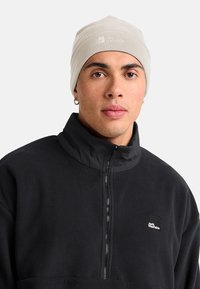 Jonge man met een beige Jack Wolfskin-beanie en een zwarte fleece-jas met rits en logo op de borst, neutrale achtergrond.