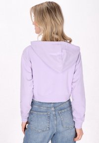 Felpa corta lavanda con polsini e orlo a coste, caratterizzata da un cappuccio foderato e una texture liscia, abbinata a jeans blu a vita alta.