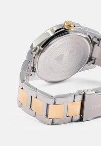 Reloj de acero inoxidable con eslabones en tono dorado, caja redonda y corona texturizada dorada. La parte trasera grabada incluye un diseño del mapa mundial.