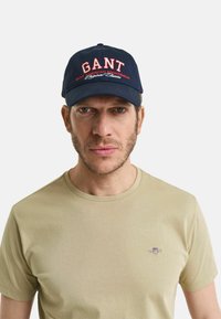 Marinblå baseballkeps med röd och vit "GANT"-logotyp och text. Mjuk tyg, rundad skärm. Ljus beige t-shirt med rund hals under.