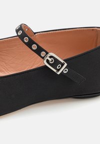 Scarpa piatta nera con tomaia in tessuto texturizzato, una cintura sottile con occhielli metallici e una fibbia argentata, e una fodera liscia beige.