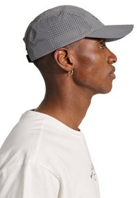 Szara czapka z perforowanymi bokami, zakrzywionym daszkiem i regulowanym paskiem z tyłu. Noszona z gładką białą koszulą. Widok z boku.