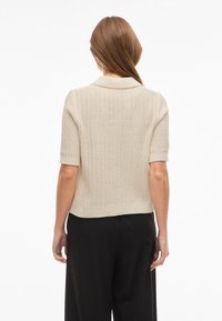 Beżowy kardigan z krótkim rękawem, wykonany z dzianiny, z kołnierzykiem, prążkowanym dołem i teksturowanym materiałem. Posiada luźny krój.