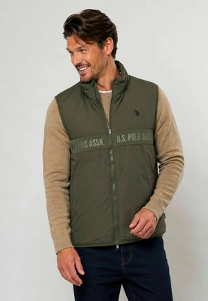 Ein Mann lächelt und trägt eine grüne ärmellose Weste mit Reißverschluss und U.S. Polo Assn.-Branding über einem beigen Pullover und dunklen Jeans.