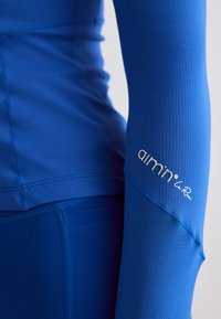 Haut de sport texturé bleu à manches longues avec logo "aimn" sur l'avant-bras et leggings bleus assortis, porté sur le torse et le bras d'une personne.