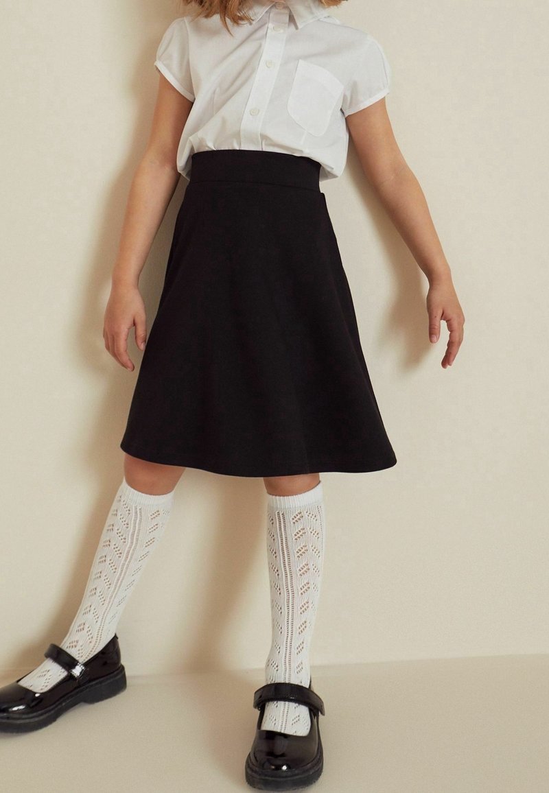 Enfant portant une chemise blanche à manches courtes boutonnée, une jupe noire longueur genoux, des chaussettes blanches montantes à motifs et des chaussures Mary Jane noires.