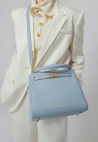 Svijetloplava kožna torba s izrađenim dizajnom, zlatnim metalnim detaljima, gornjom ručkom i odvojivim remenom za rame. Teksturirana površina i detalji s logotipom.