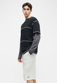 Jeune homme portant un t-shirt à manches longues superposé noir et gris et un short gris clair arrivant aux genoux, debout devant un fond blanc.