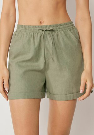Frau trägt locker sitzende Leinen-Shorts in Olivgrün mit elastischem Bund, Kordelzug, seitlichen Taschen und umgeschlagenen Säumen.