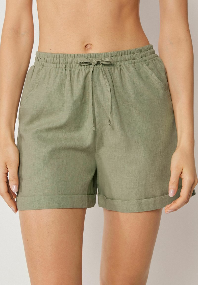 Frau trägt locker sitzende Leinen-Shorts in Olivgrün mit elastischem Bund, Kordelzug, seitlichen Taschen und umgeschlagenen Säumen.
