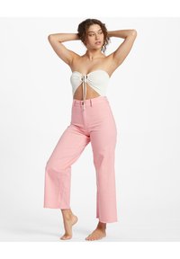 Donna in posa a piedi nudi, indossando jeans a vita alta e a gamba larga di un leggero rosa e un top corto bianco senza spalline con dettagli di ritaglio e nodo.