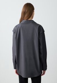 Chemise surdimensionnée gris foncé avec une texture lisse, dotée d'un col classique et de manches retroussées, l'ourlet étant plus long à l'arrière.