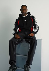 Uomo seduto su un armadietto grigio, indossa una felpa nera Adidas con accenti rossi, jeans neri larghi e scarpe nere con design bianchi.