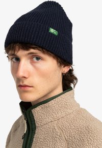 Gorro de punto acanalado azul marino con parche de logo verde; el modelo también lleva una chaqueta de forro polar beige con ribete verde y un cuello alto.