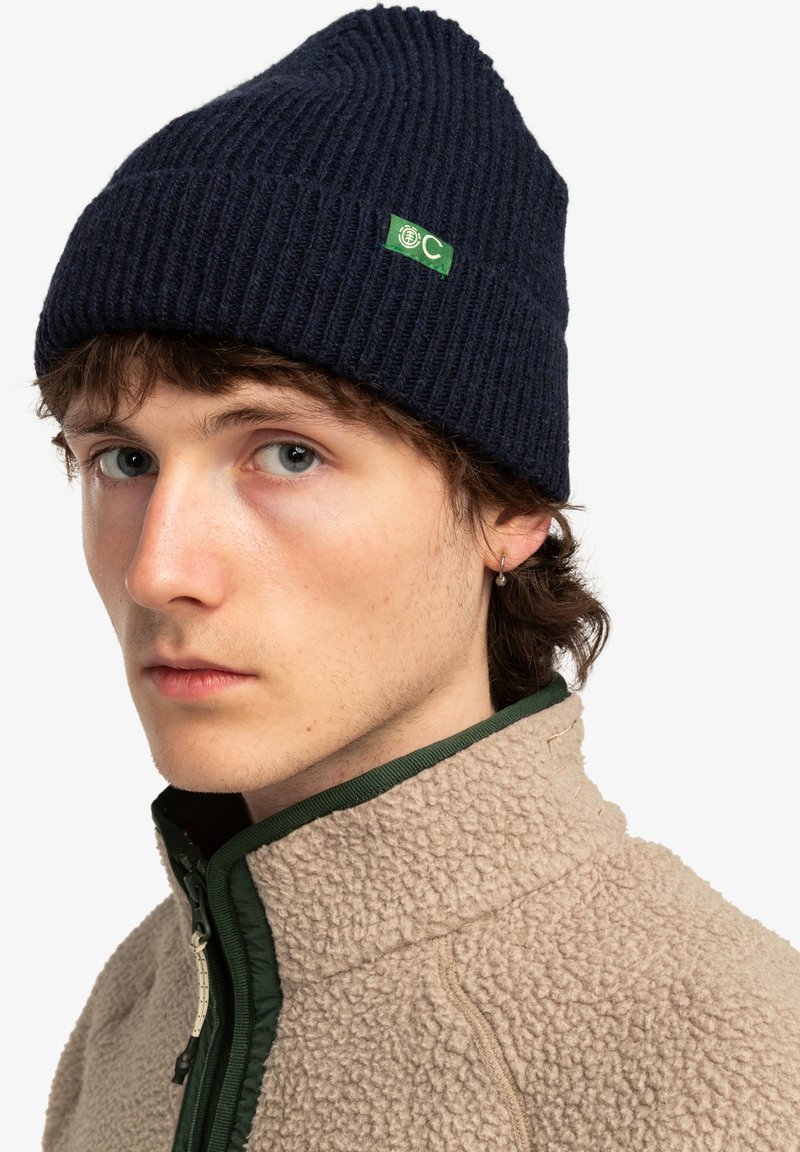 Gorro de punto acanalado azul marino con parche de logo verde; el modelo también lleva una chaqueta de forro polar beige con ribete verde y un cuello alto.