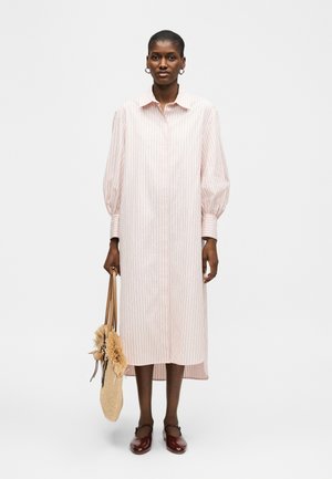 Kvinne iført en lang, lyserosa stripete skjortekjole, holder en farget vevd handleveske med frynser, og har mørkebrune Mary Jane-sko på.