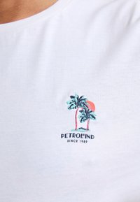 Bílý bavlněný tričko s vyšívaným designem dvou palem a slunce, pod nimi je modrý text "PETROLIND" a "SINCE 1989".