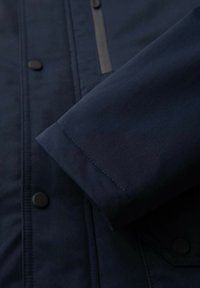 Veste bleu marine à texture lisse, avec une manchette à boutons et des coutures visibles. Comprend une poche zippée à l'avant.