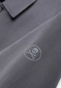 Tejido gris con un acabado mate; presenta un logo redondo que muestra una calavera y huesos cruzados en un tono más claro en la parte inferior derecha.