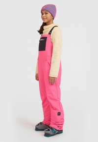 Felle roze sneeuwoverall met zwarte accenten, een crème top met fleece-voering, en grijze skibotten met blauwe details, gedragen door een persoon die zijwaarts staat.