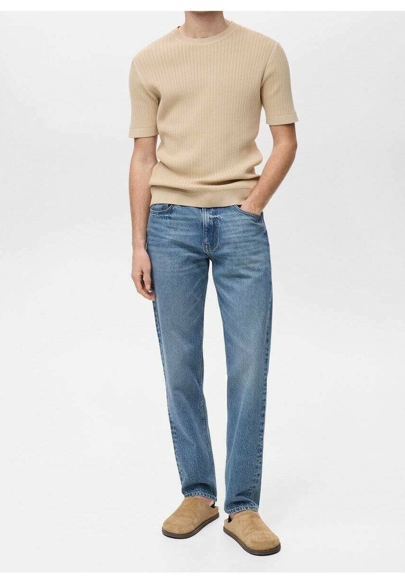 Homme portant un pull côtelé à manches courtes beige, un jean droit bleu et des chaussures à enfiler marron clair, debout avec une main dans la poche.