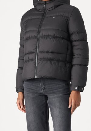 Persoon draagt een zwarte donsjas met een ritssluiting aan de voorkant en drukknopen aan de manchetten, gecombineerd met donkergrijze spijkerbroeken, staand tegen een effen achtergrond.