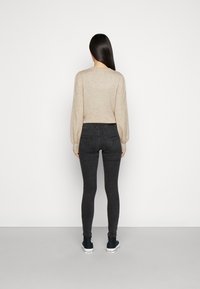 Suéter de punto beige con mangas abullonadas, combinado con jeans ajustados negros y zapatillas negras. El fondo liso realza los detalles de la ropa.